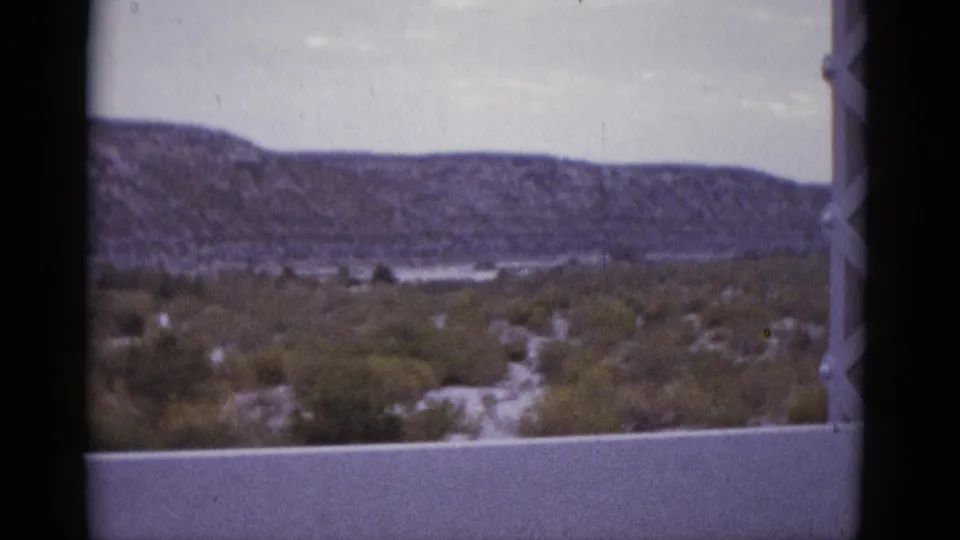 1952 8mm frame of a Texas desert valley, flat mesa prominent in mid-background, with low shrubs and a white overcast sky.