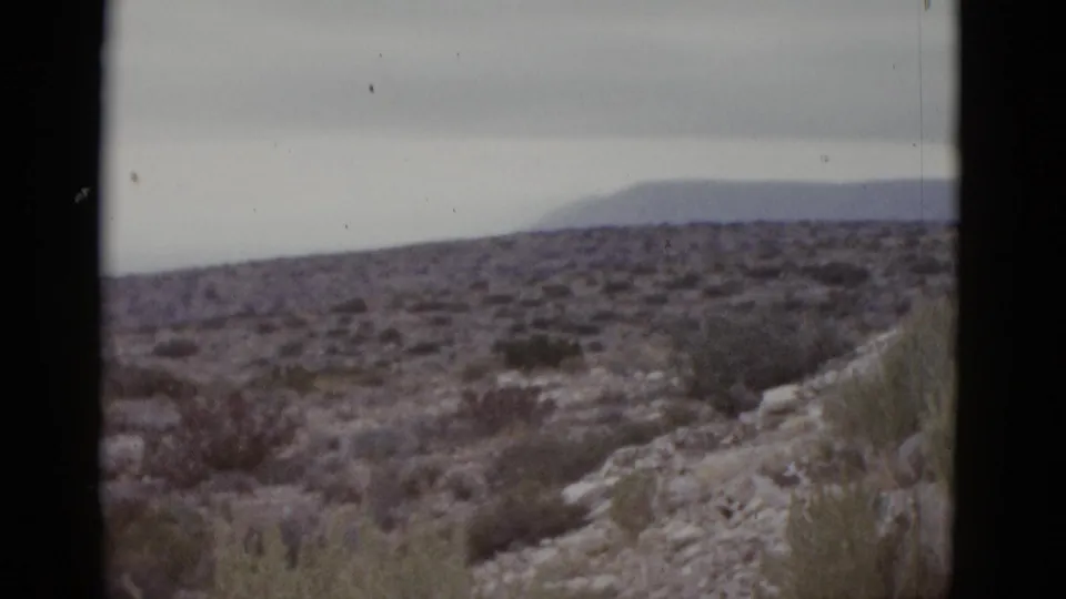 1952 8mm frame showing a rocky desert ridge in the foreground, arid scrubland, and a distant mountain range on the horizon under a flat overcast sky, likely Texas.