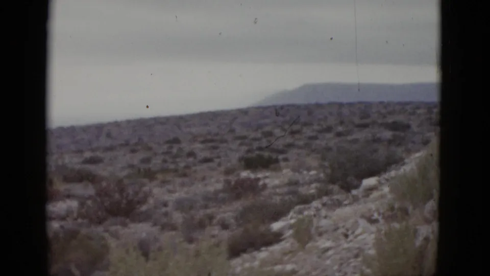 8mm frame from a 1952 Texas road trip showing a rocky arid hillside with sparse desert shrubs and a hazy mountain ridge on the horizon under flat gray skies.