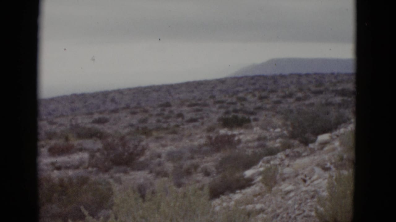 Arid desert slope with rocky outcrops and sparse scrub, distant mountain ridge against overcast sky; near-identical framing to opening frame.