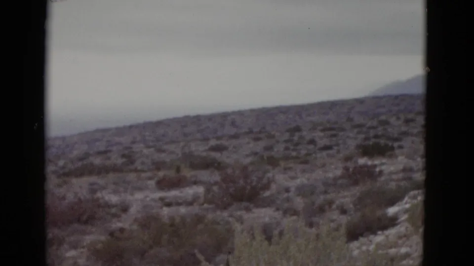 Closing 8mm frame from 1952 Texas desert pan, scrubland in foreground and a mountain ridge at the right horizon under flat gray skies.