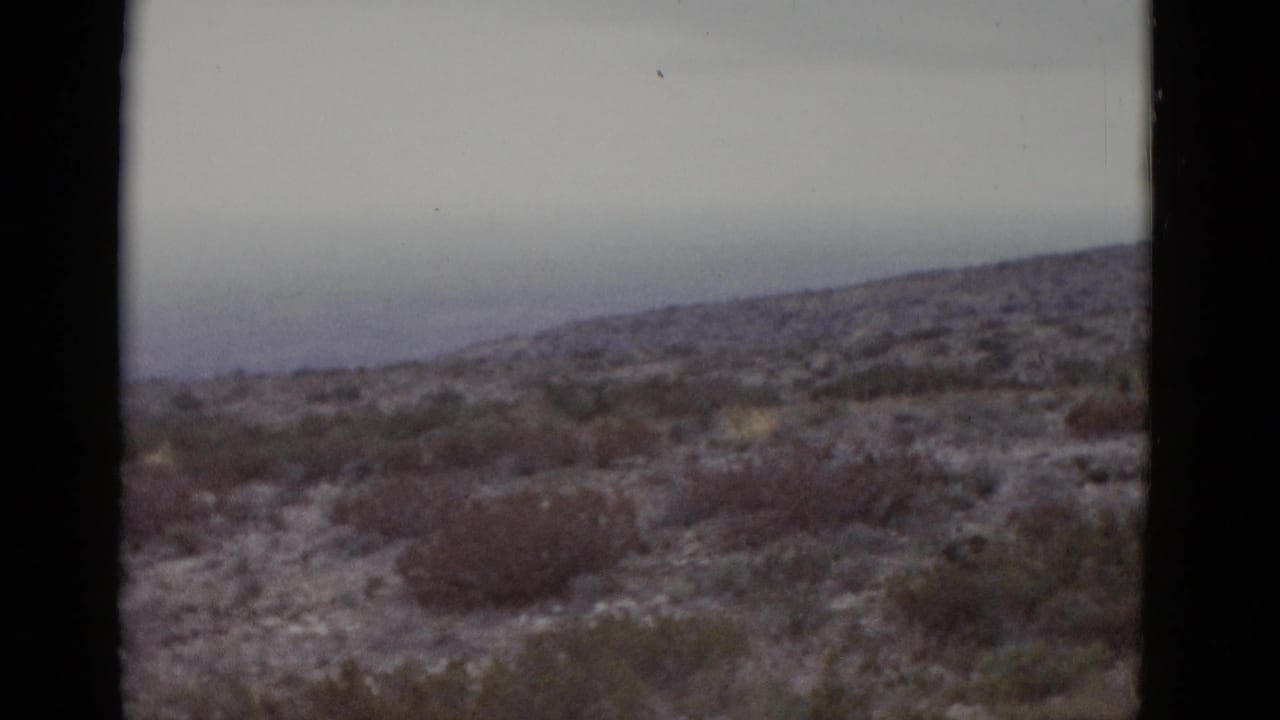 1952 8mm home movie frame of Texas arid hills — rocky scrubby terrain under an overcast sky with visible film dust artifacts.