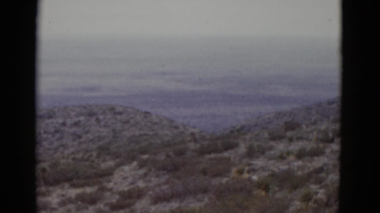 Same desert hill from a similar angle, sparse low scrub on an arid slope, overcast sky, 8mm frame borders visible.