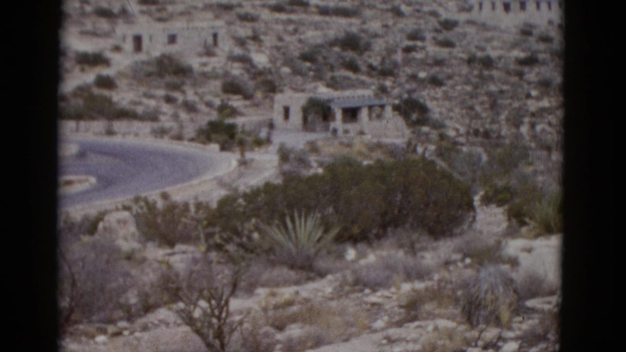 Stone building with arched arcade on a desert hillside, winding road below, agave plants in foreground.