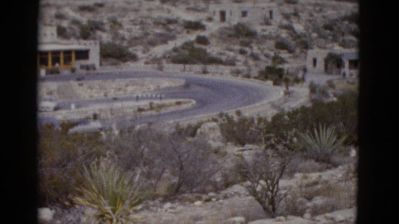 Mid-pan 1950s 8mm frame over Utah desert showing curved road, rocky terrain, agave plants, and stone structure nestled in upper hillside