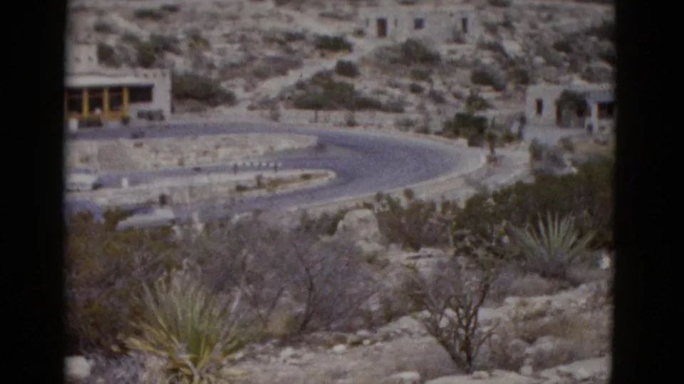 Mid-pan 1950s 8mm frame over Utah desert showing curved road, rocky terrain, agave plants, and stone structure nestled in upper hillside