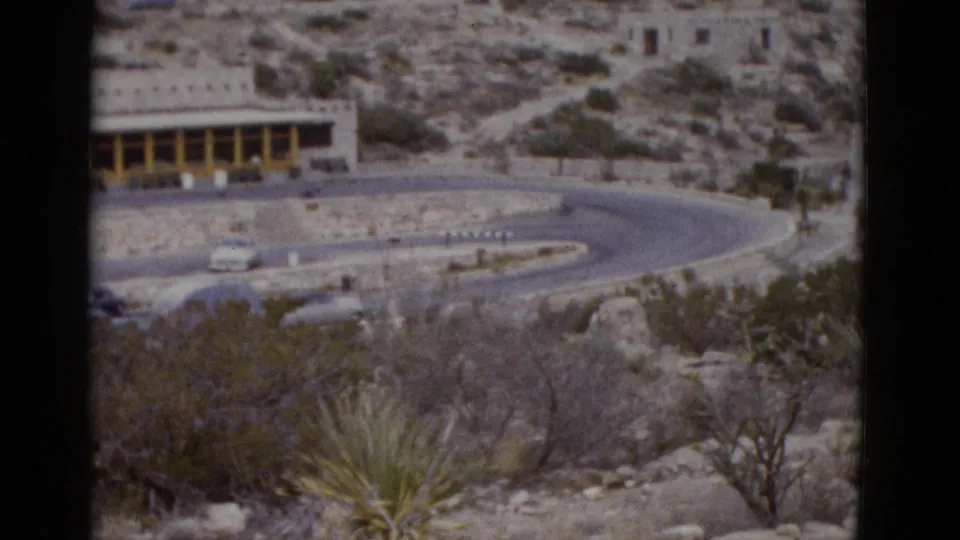 1950s 8mm footage revealing a long, low stone roadside building with yellow-painted colonnade and vintage automobiles parked outside, surrounded by desert scrub