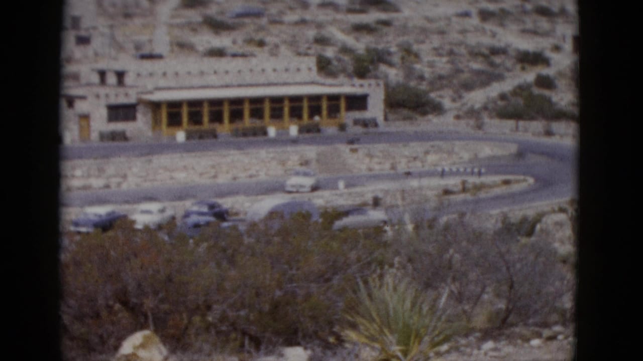 Wide elevated shot of a stone building with yellow colonnade in a Utah desert landscape, 1950s cars visible in foreground lot.