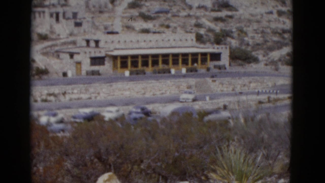 Elevated pan view of a stone building with yellow beam colonnade set against a rocky desert hillside, 1950s cars in parking area.