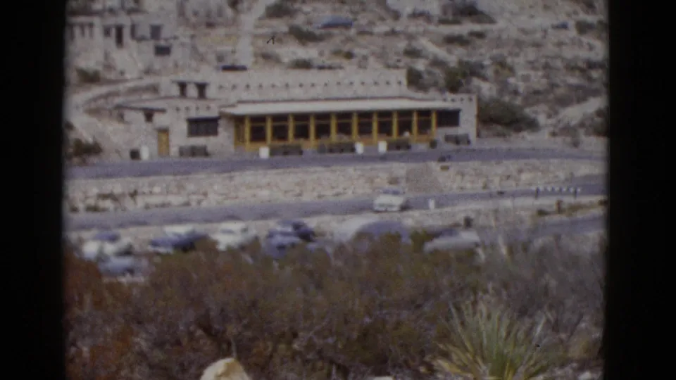 Pan frame showing a low-profile stone building with yellow colonnade in Utah arid landscape, circa 1952, with vintage American cars and desert vegetation visible.