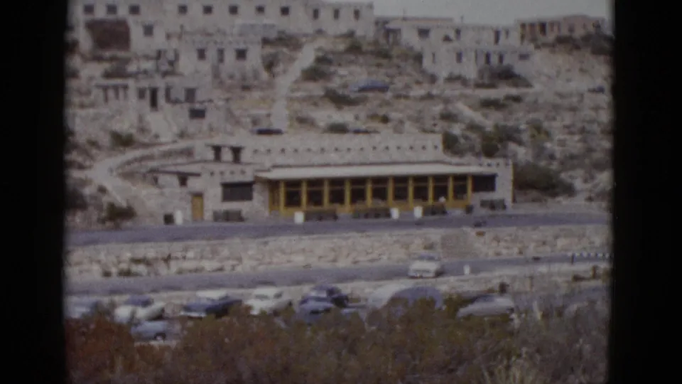 Late pan frame of a mid-century stone building in Utah desert, circa 1952, showing curved road, 1950s cars, desert scrub, and rocky hillside terrain.