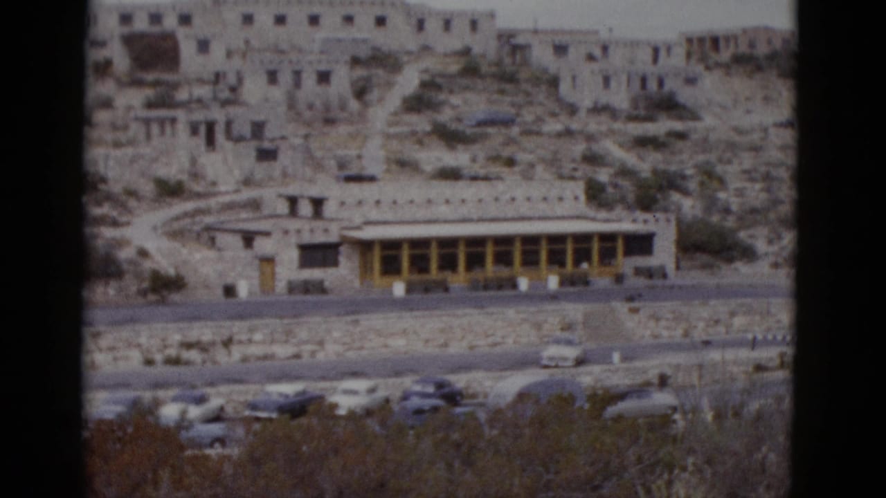 Stone lodge complex on rocky desert hillside with vintage 1950s cars parked below and yellow-trimmed lower facade.