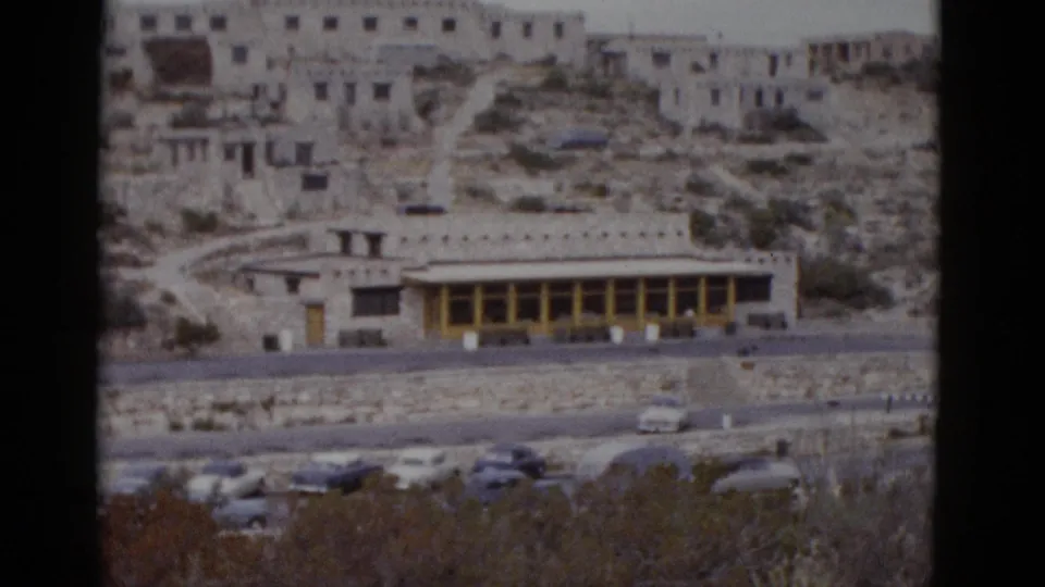 Multi-storey stone lodge with yellow-trimmed facade and vintage automobiles in parking area, American Southwest desert landscape, circa 1952 8mm footage.