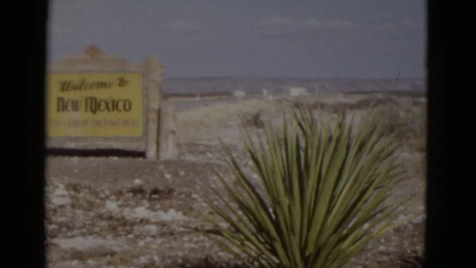 Yellow 'Welcome to New Mexico' state welcome sign on wooden posts with yucca plant at right, flat desert and pale sky; 1952 8mm road-trip footage.