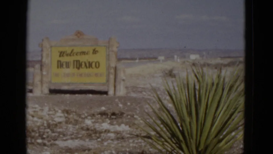8mm frame from a 1952 Southwest road trip: New Mexico state-line welcome sign on stone pillars, yucca plant foreground, open desert stretching to the horizon.