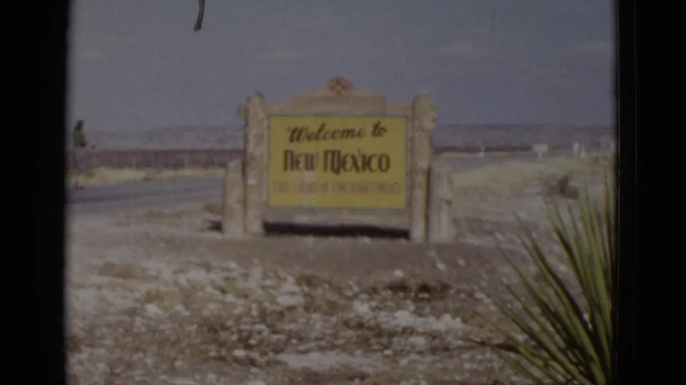 8mm frame of New Mexico border welcome sign from a shifted viewpoint, c. 1952; a small figure appears at the far left margin and warrants review.