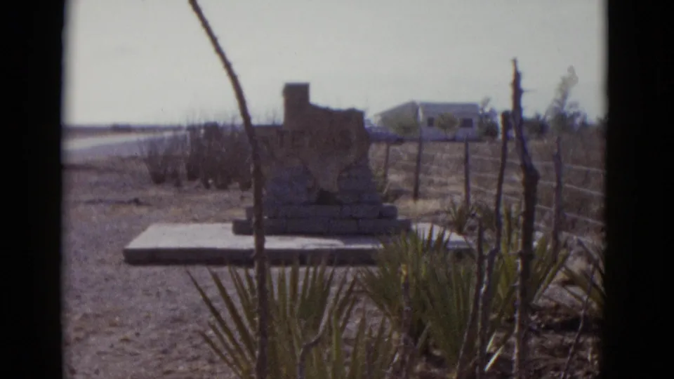 8mm film frame, circa 1952, showing a Texas-shaped roadside stone monument in an arid Southwest landscape with yucca plants and a distant vehicle.