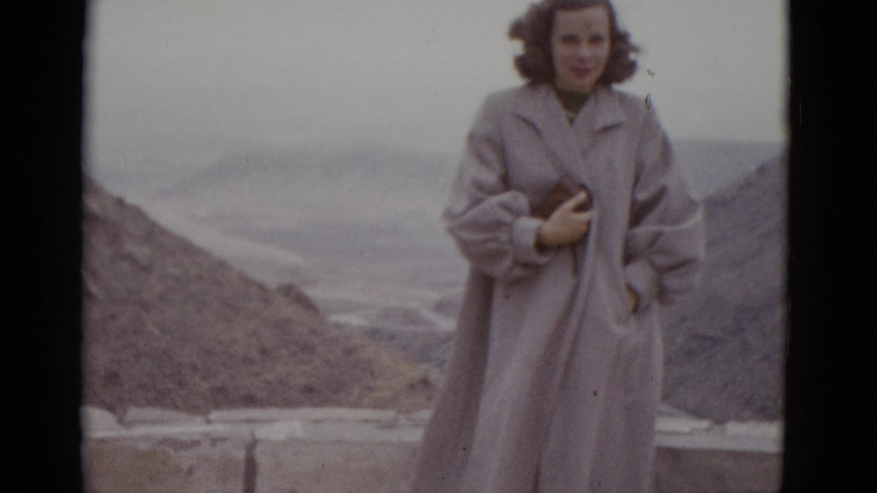 Young woman in long pale coat stands at a desert roadside overlook, dry rocky hills behind her, overcast sky above.