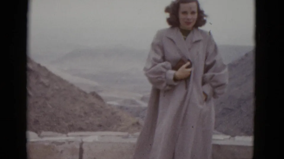 Young woman in full-length pale coat poses at a Utah desert overlook, 1952; bare rocky hills and flat overcast sky visible behind a low stone parapet.