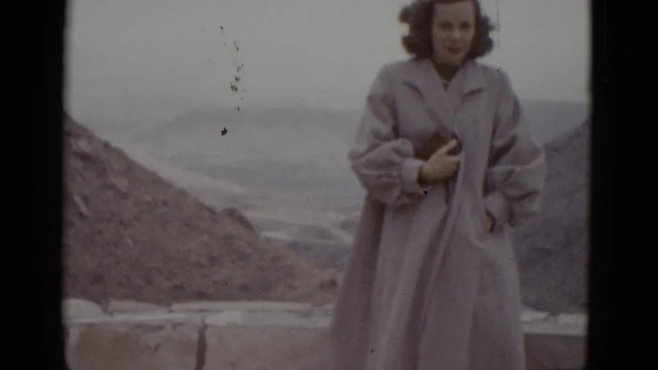 Young woman in full-length pale coat at a Utah desert roadside stop, 1952; film dust artifacts visible upper right, dry hills and overcast sky behind.