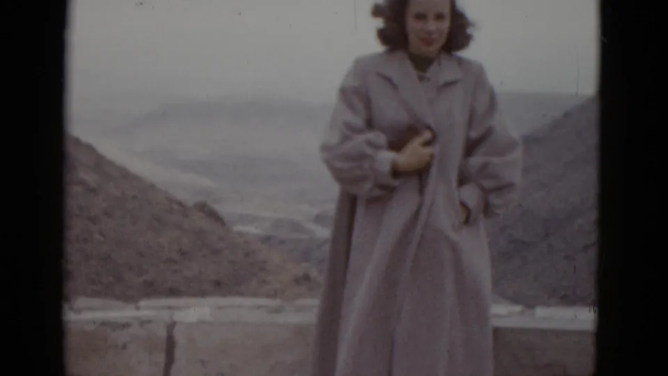 Young woman in full-length pale coat at Utah desert roadside stop, 1952; subtle expression change visible, arid landscape behind low stone parapet.