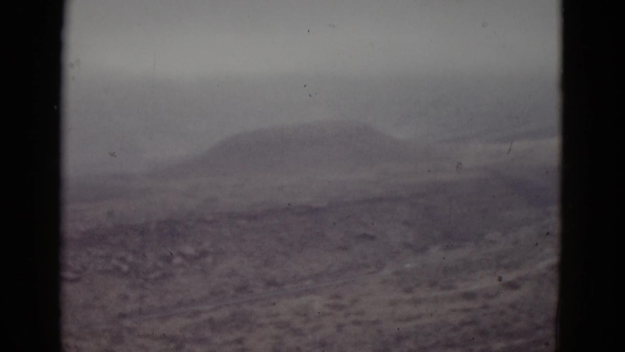 1950s 8mm home movie frame showing a low rounded mountain peak over barren high-desert terrain in Utah, with soft focus and cool purple-gray color cast.