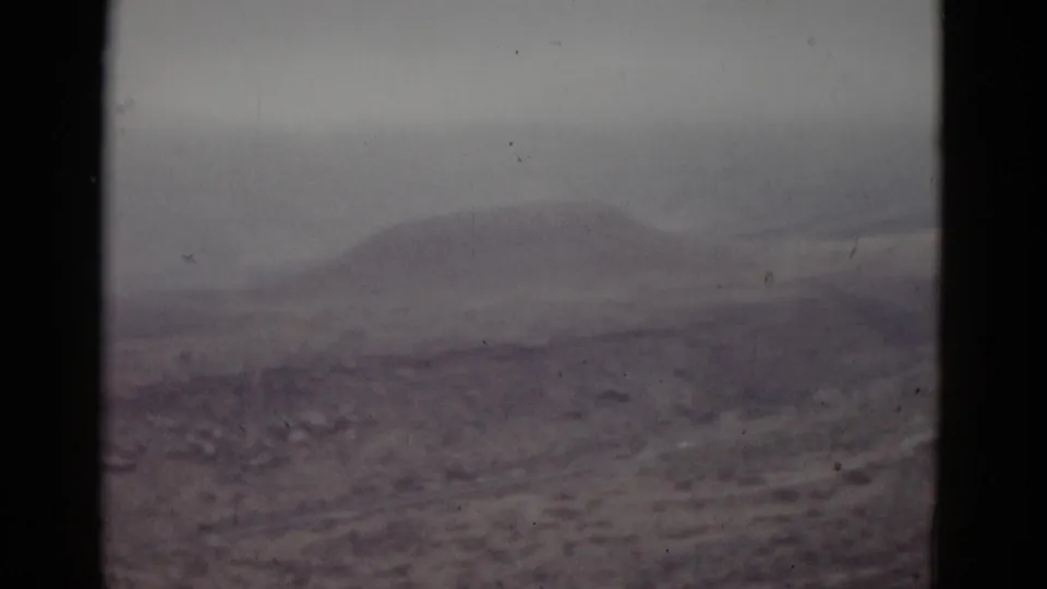 1950s 8mm home movie frame showing a low rounded mountain peak over barren high-desert terrain in Utah, with soft focus and cool purple-gray color cast.