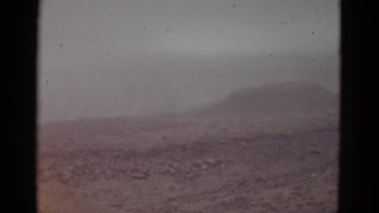Same hazy mountain silhouette; foreground terrain shows a warmer reddish-pink color shift compared to the opening frame.