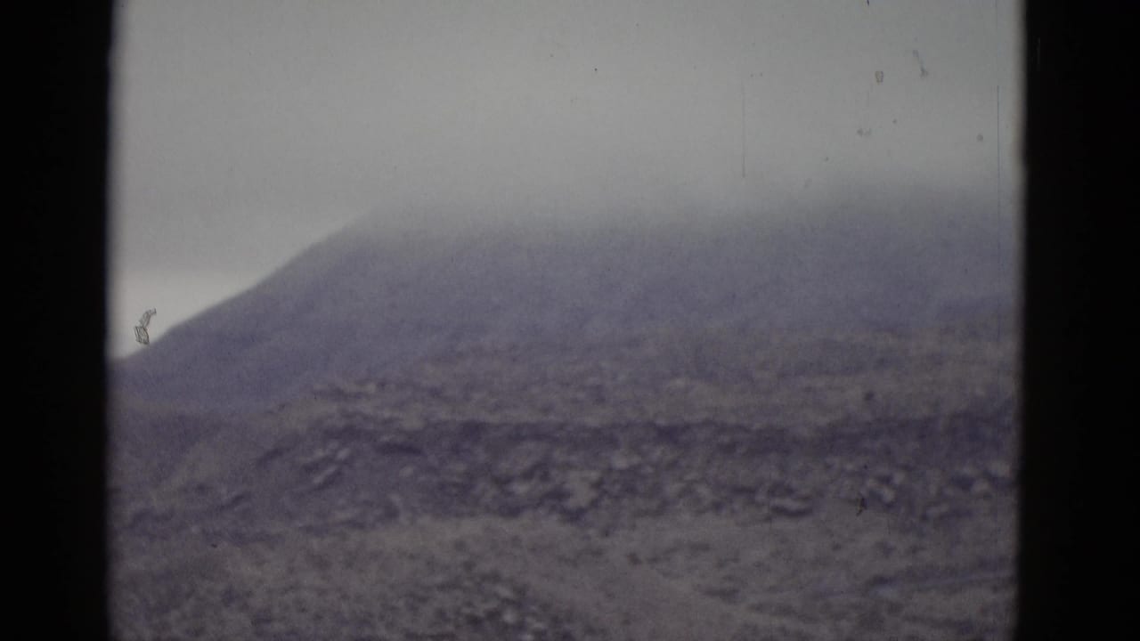 Bare conical mountain peak against overcast gray sky with rocky scree slopes, filmed on 8mm color film.