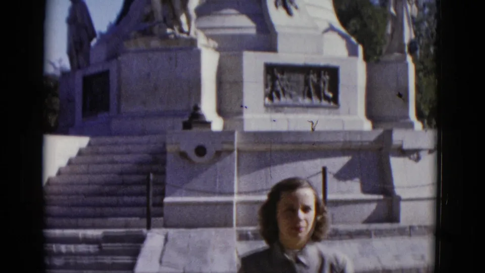 Wide shot of woman at the base of a large white stone commemorative monument in Utah, 1952; bas-relief panels, stone piers, and trees visible behind her.