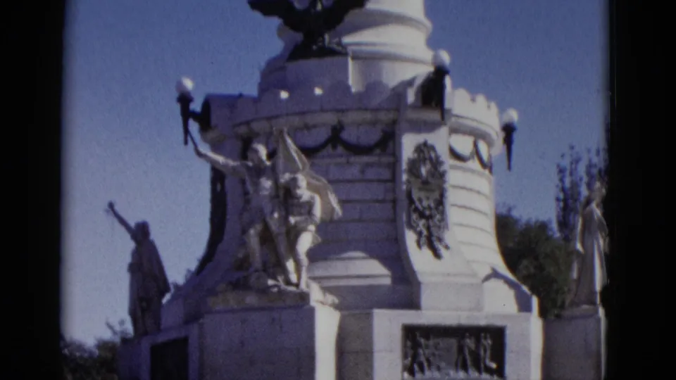 8mm frame from 1952: white civic monument mid-tilt with sculptural figures, crenellated battlements, lamp fixtures, and trees visible against blue sky.
