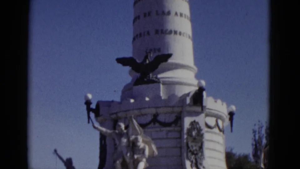 8mm frame from 1952: monument column apex with spread-wing eagle and two partial lines of Spanish text visible on the pillar, camera at top of upward tilt.