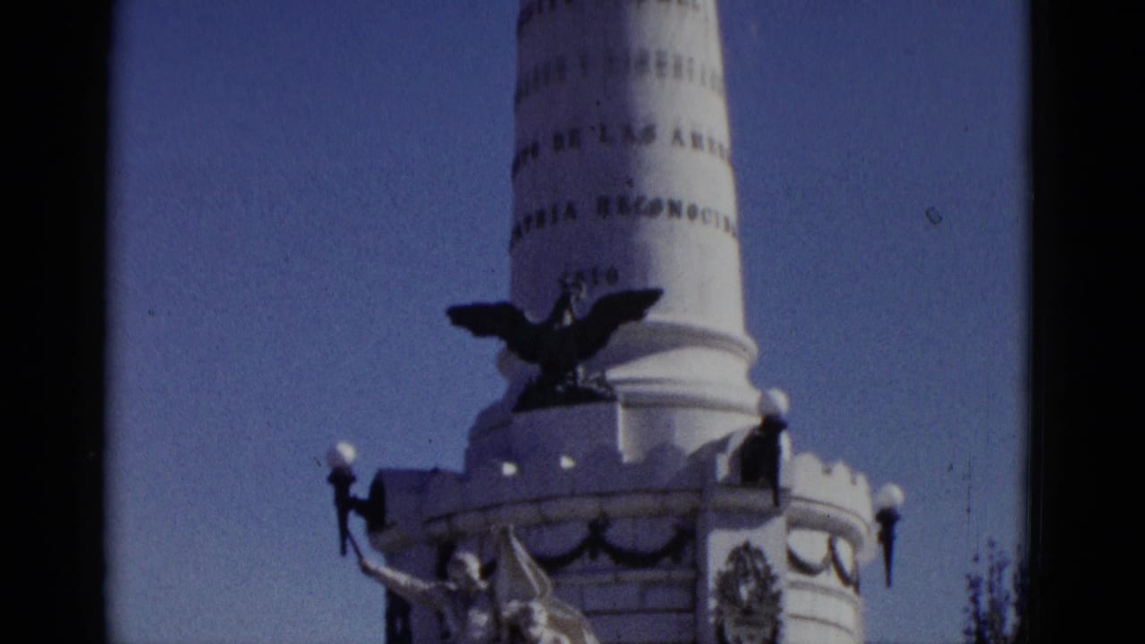 Low-angle 8mm view of a tall white monument column with Spanish inscriptions and an eagle sculpture against a blue sky.