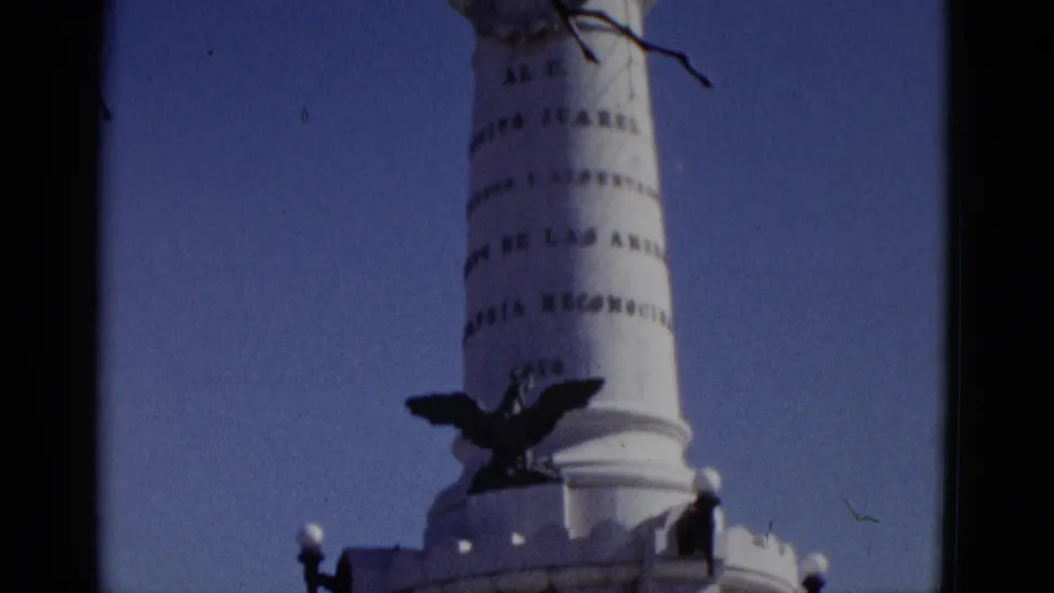 Mid-tilt 8mm frame of a white memorial column bearing Spanish-language inscriptions and a spread-eagle sculpture, from a 1952 home movie attributed to Utah.