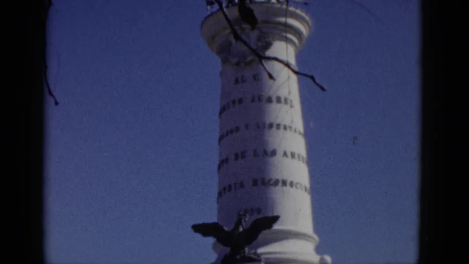 1952 8mm home-movie frame of a white column with eagle, inscriptions appearing to include the name 'JUAREZ,' and tree branches framing the shot.