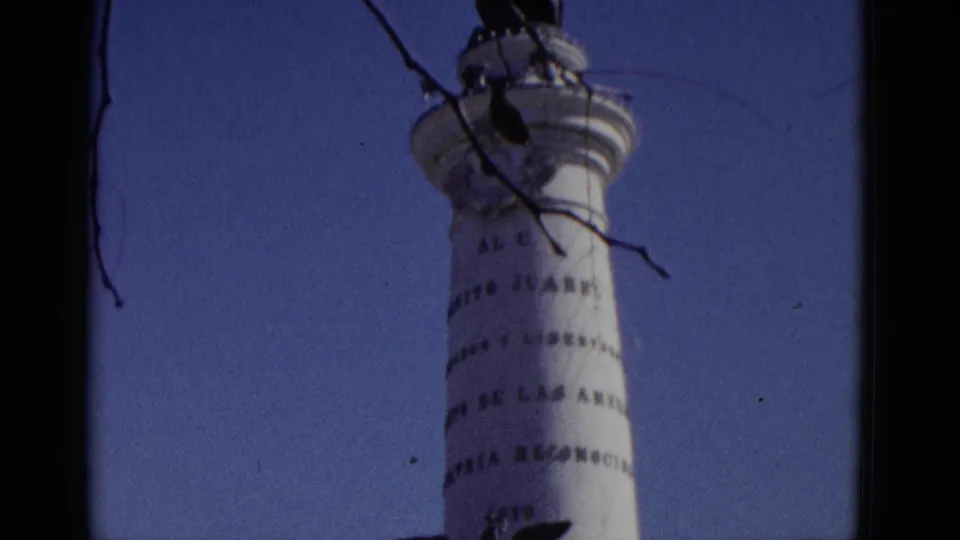 Final frame of 1952 8mm tilt shot; white commemorative column with spread-eagle and partial Spanish inscriptions fills the frame against a clear blue sky.