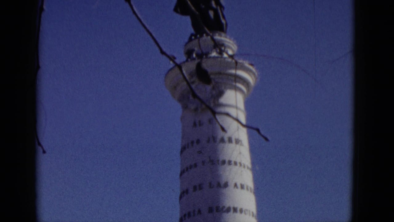 1952 8mm home-movie upward angle of a white commemorative column bearing a partially legible Spanish inscription, dark statue silhouetted at top, bare winter branches against blue sky.