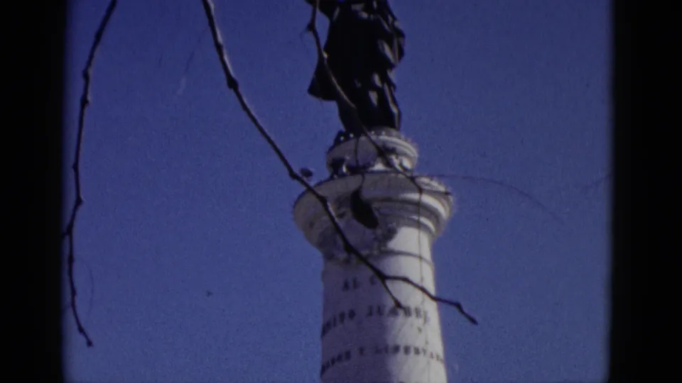 1952 home-movie frame of a white commemorative column showing legible fragments of a Spanish dedication text, dark figure at top, bare branches against a deep blue sky.