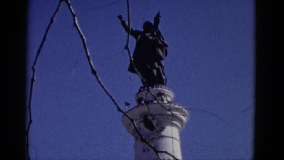 1952 home-movie low-angle shot of a dark figurative statue with outstretched arms crowning a white monument column, framed by bare tree branches against a saturated blue sky.