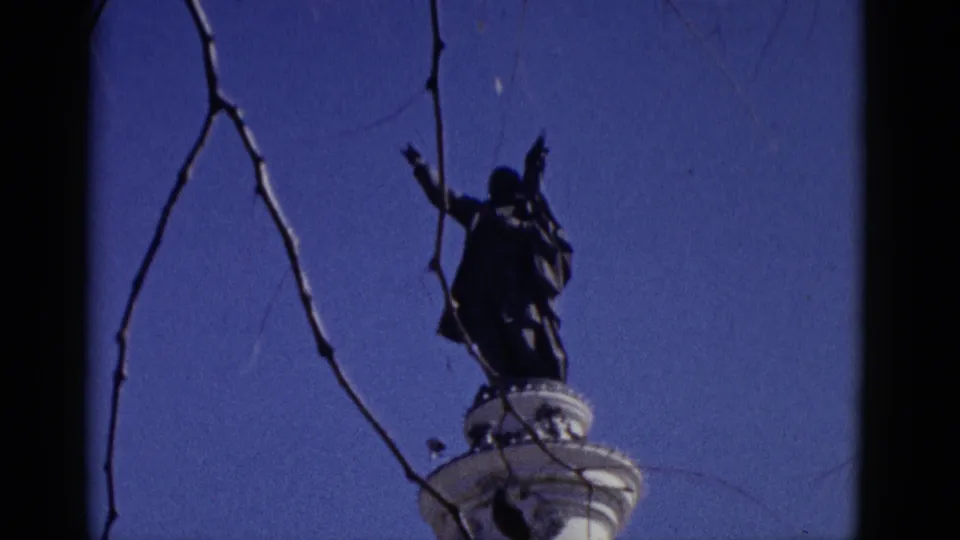 Final 1952 home-movie travelogue frame: dark figurative statue with one arm extended atop a white column, framed by bare branches against a clear, saturated blue sky.