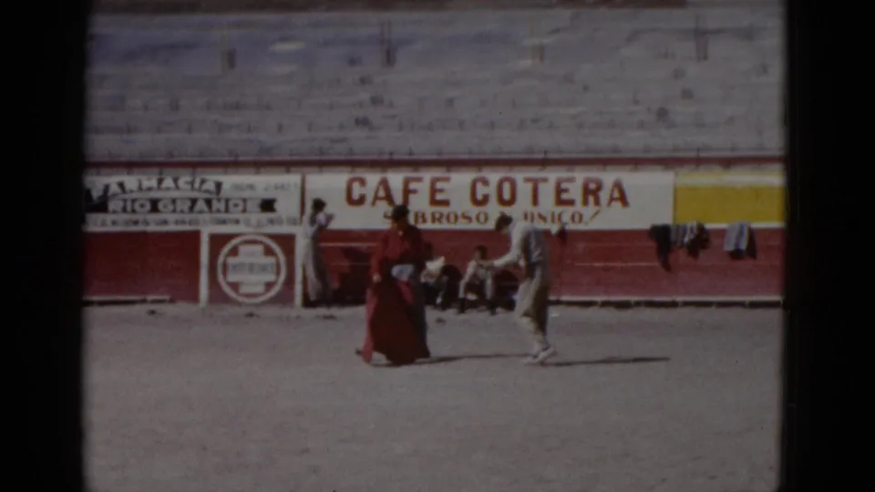 Two practitioners in what appears to be a 1952 Baja California bullring; one in red attire, one in white; Cafe Cotera and other signs on arena barrier.