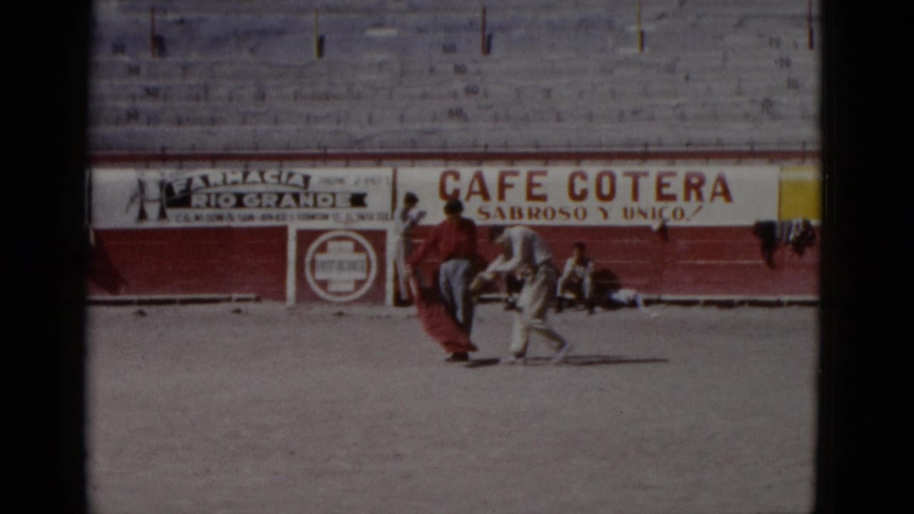 Two people rehearse cape work on the sand of a Baja California bullring, circa 1952; period advertisements for Cafe Cotera and Farmacia Rio Grande are legible on the red barrera wall.