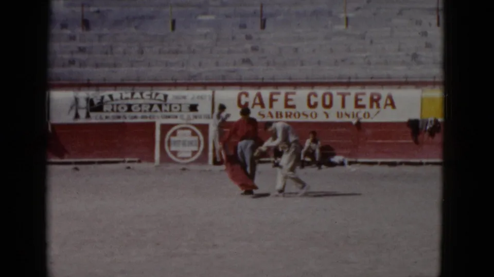 Two people rehearse cape work on the sand of a Baja California bullring, circa 1952; period advertisements for Cafe Cotera and Farmacia Rio Grande are legible on the red barrera wall.