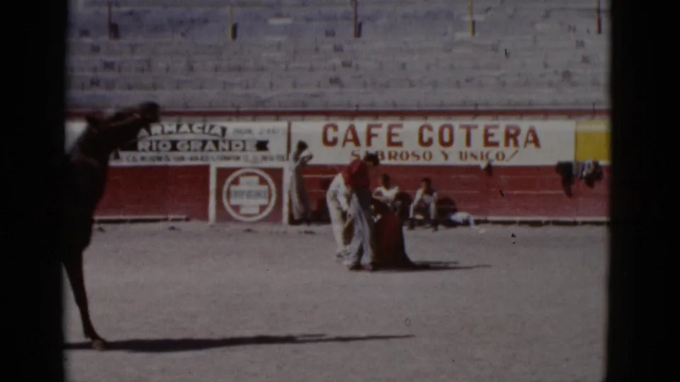 A practitioner in light-colored clothing handles a cape near the barrera of an empty bullring, circa 1952, with Cafe Cotera and Farmacia Rio Grande advertisement banners visible behind.