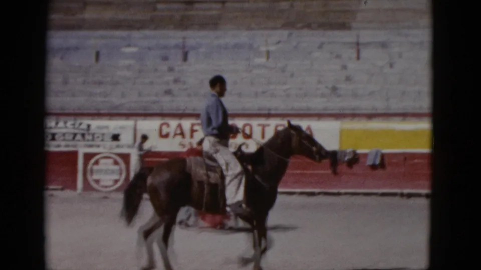 A mounted rider on a dark brown horse moves across a sunlit bullring in what appears to be Baja California, circa 1952, with period commercial banners on the barrier wall behind.