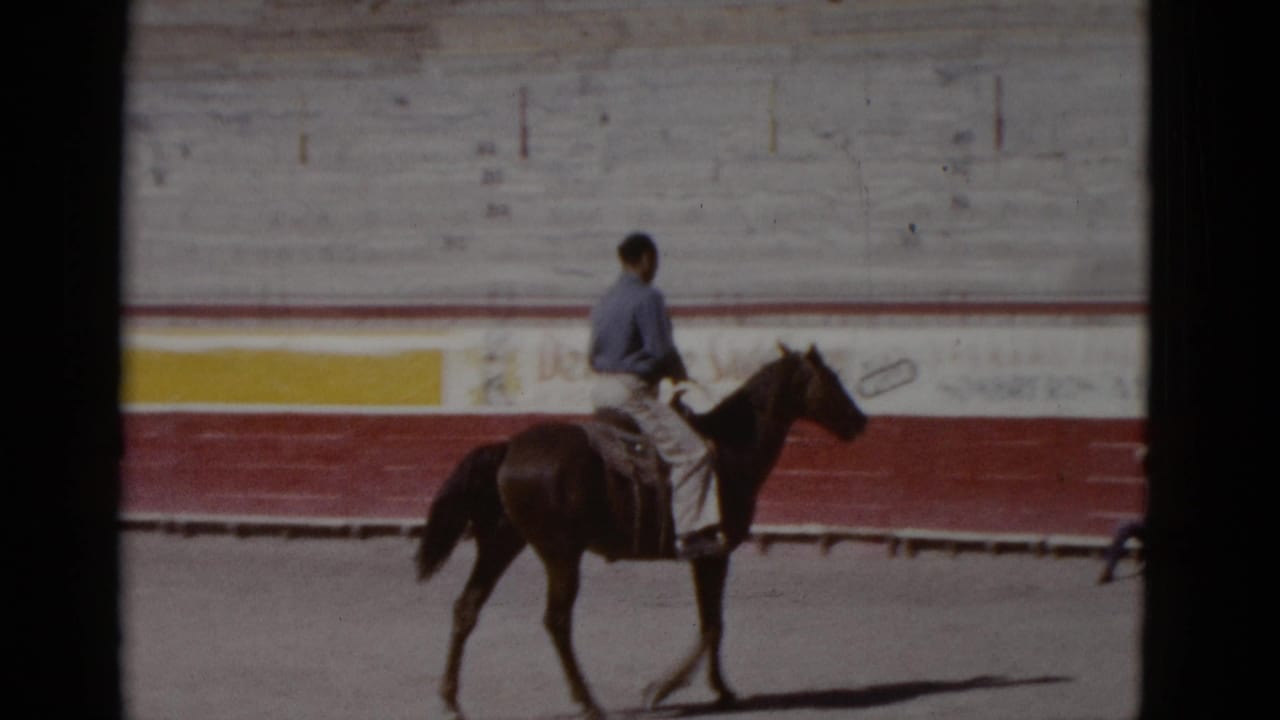 Rider on a dark horse continues trotting along the arena wall, partially legible Spanish-language advertisements painted in red and yellow behind him.