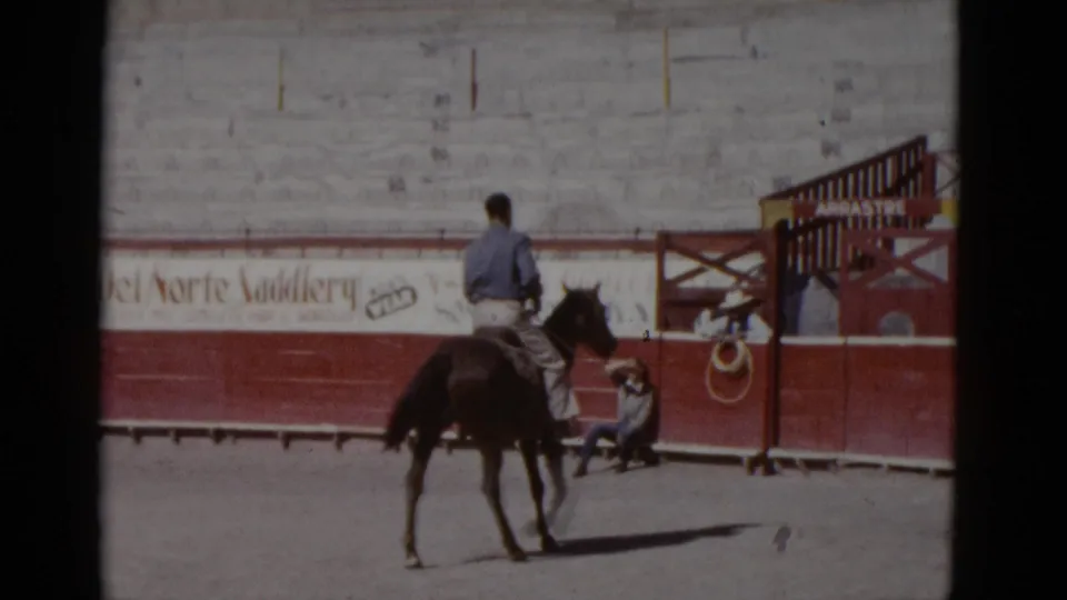Horseman seen from behind approaching a red wooden gate structure in the arena; 'del Norte Saddlery' advertisement on the bleacher wall and a yellow sign on the gate — Baja California, circa 1952.