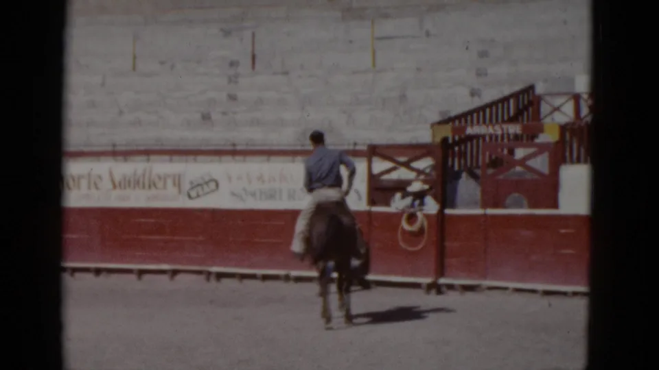 The horseman slows near the arena gate in this closing frame; 'del Norte Saddlery' advertisement remains on the bleacher wall and a yellow sign marks the wooden gate — Baja California, circa 1952.