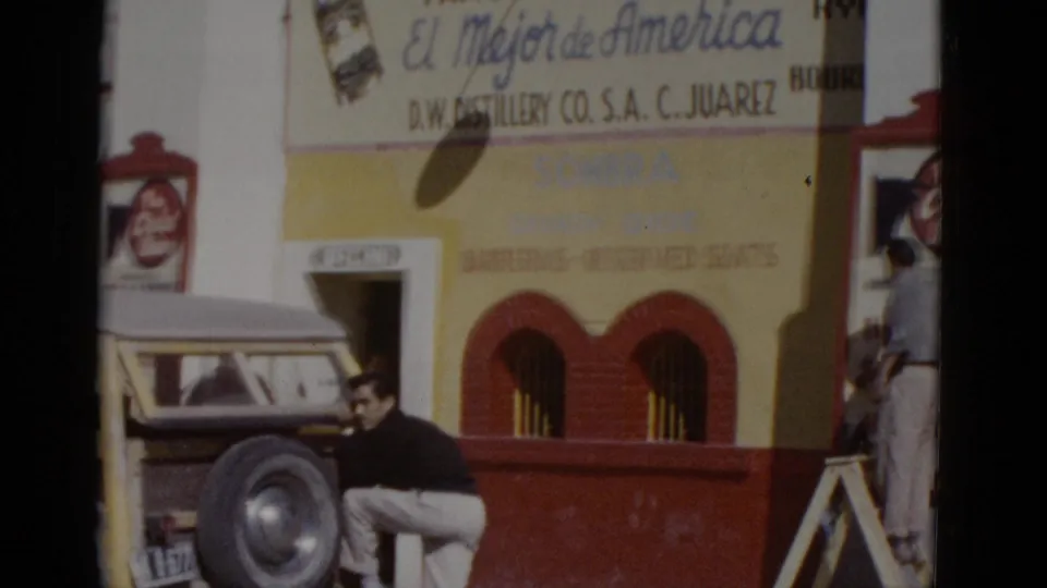 Man leaning against a vintage vehicle outside what may be a bullring or commercial building in northern Mexico, c.1952, with 'El Mejor de America' whiskey sign above.