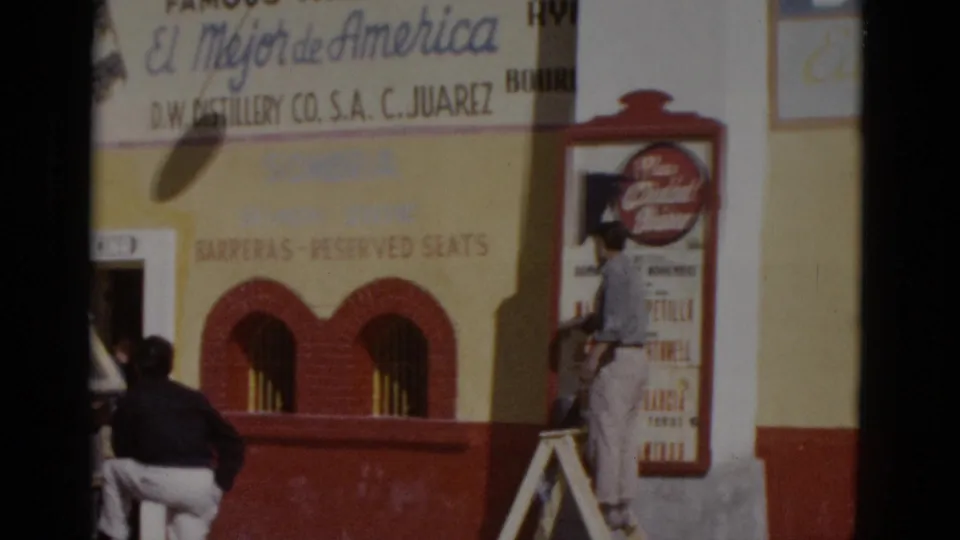 Worker on ladder tending to a 'Plaza Ciudad Juarez' arena poster on a building facade with 'El Mejor de America' commercial signage above, northern Mexico, c.1952.